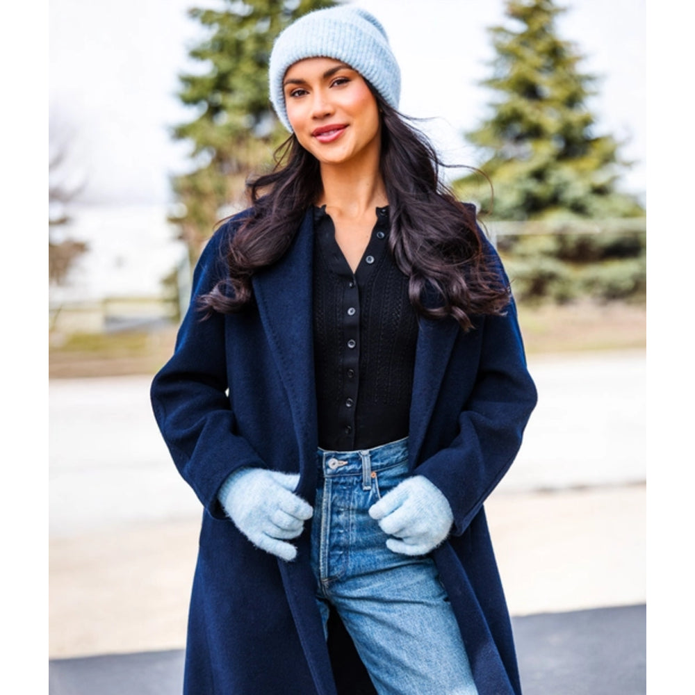 A woman wearing a long navy blue coat, black button-up top, light blue jeans, a light blue knit beanie, and matching light blue gloves, standing outdoors with blurred evergreen trees in the background.