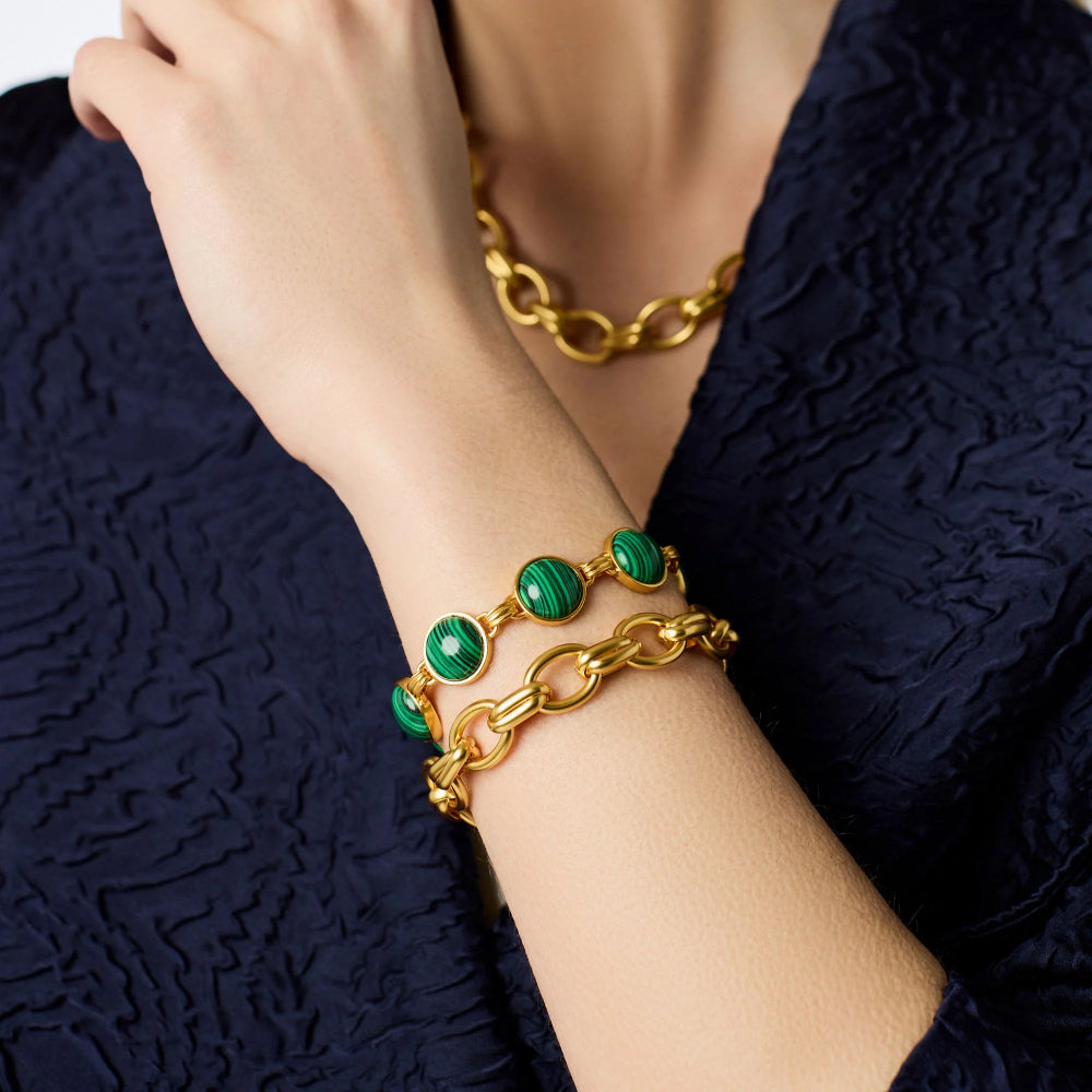A close-up of a person's wrist wearing a stack of gold bracelets, including a chain bracelet and a bracelet with green stone beads, along with a matching gold chain necklace. The person is wearing a textured navy blue top.