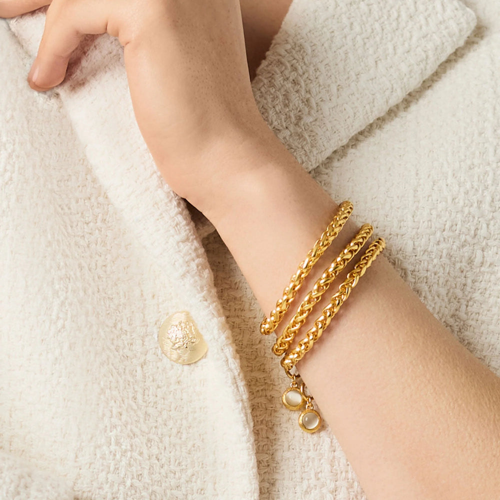 A close-up of a person's wrist wearing three stacked gold chain bracelets with small pearl-like bead accents, resting on a textured cream-colored knit garment.