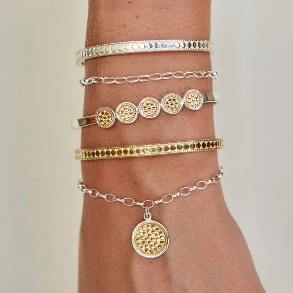 A close-up of a person's wrist wearing five stacked bracelets: two silver bangles, two gold bangles with circular medallion details, and one silver chain bracelet with a round textured gold pendant.