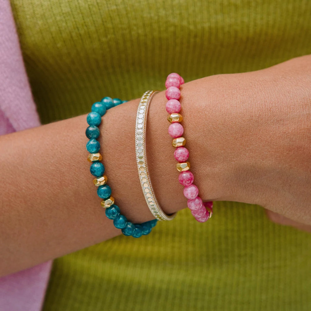 Three beaded bracelets worn on a person's wrist: a turquoise-blue beaded bracelet with gold spacer beads, a silver-toned pavé bracelet, and a pink beaded bracelet with gold spacer beads. The background shows a ribbed olive green top and a pink fabric edge.