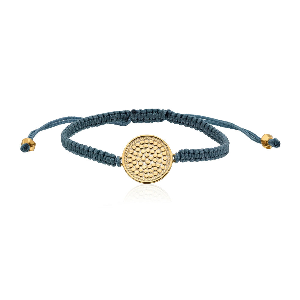 A blue grey woven  bracelet with a textured gold circular disc charm in the center and small gold sliding beads at the ends