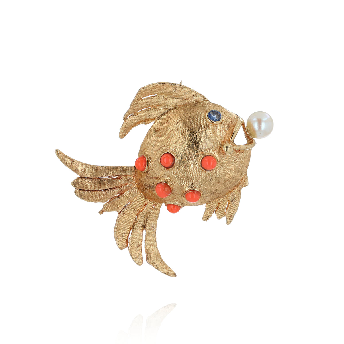 A gold fish-shaped brooch featuring coral stones on its body, a small blue sapphire for an eye, and a white pearl protruding from its mouth, set against a plain white background.