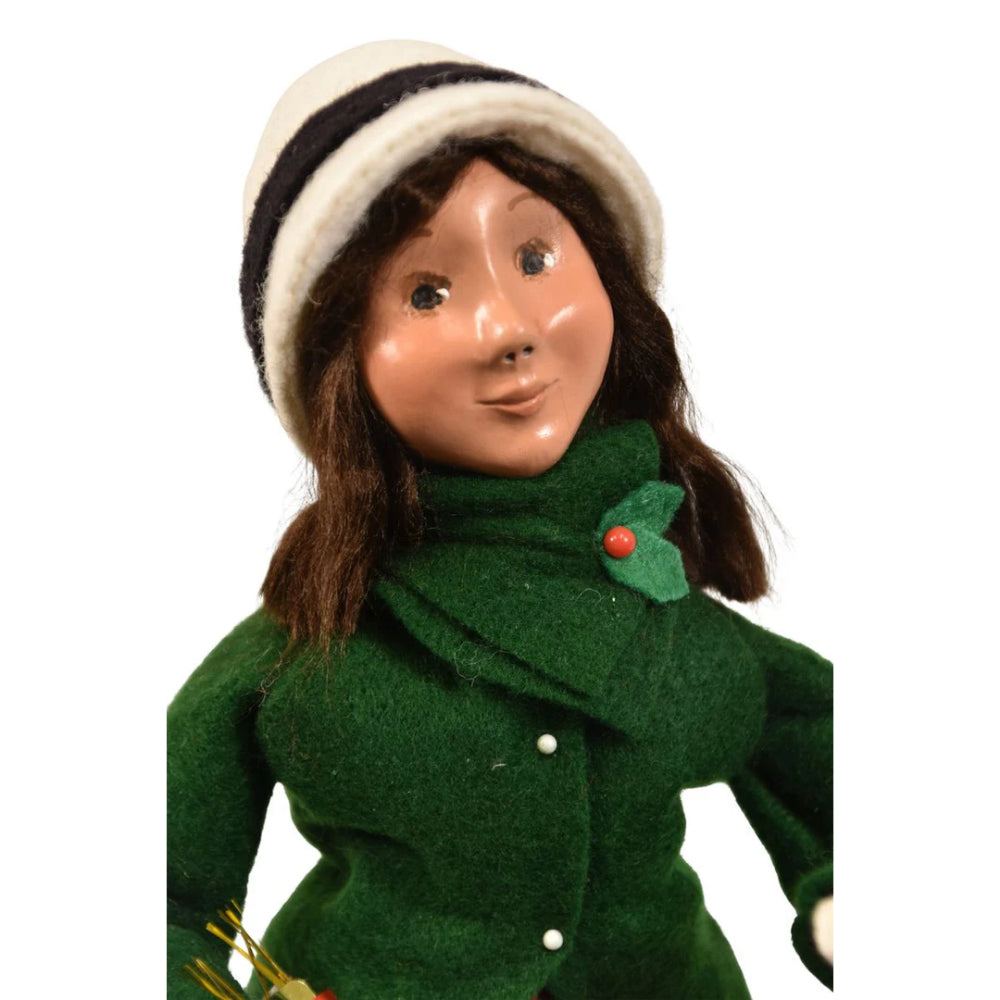 A close up photo of a figurine of a caroling girl wearing a green coat and red plaid skirt on holding a nutcracker ornament and holiday popper on a white background