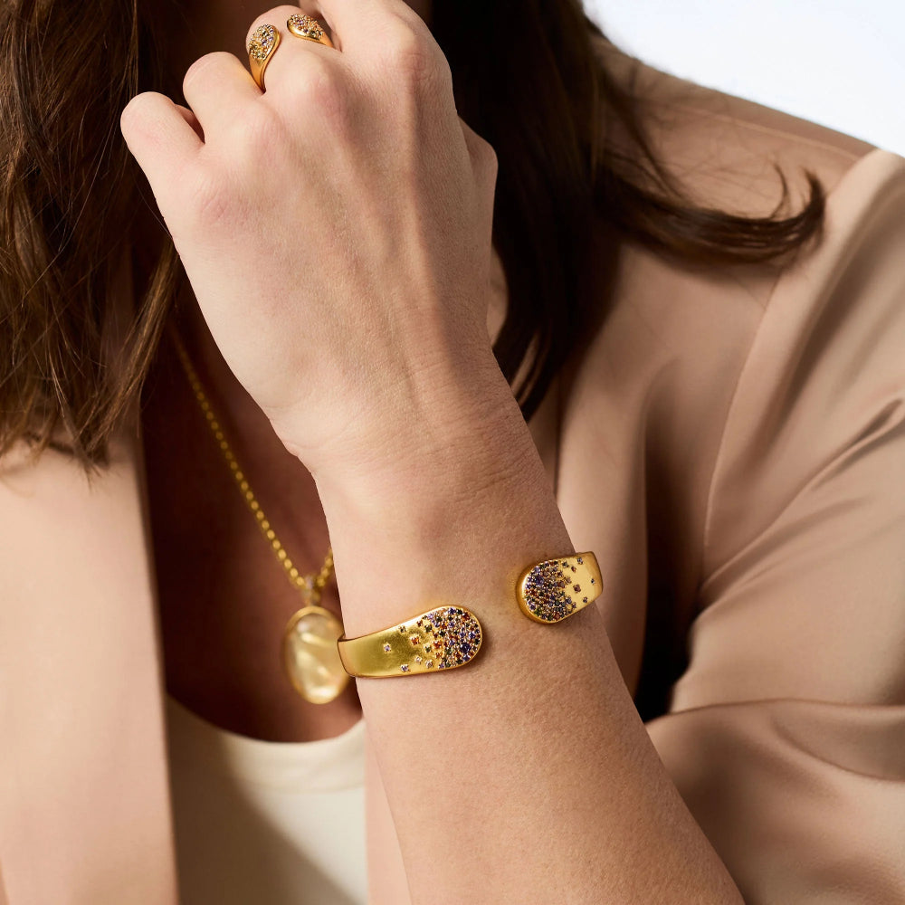 A close-up lifestyle shot of a woman wearing a gold cuff bracelet with colorful enamel dots, along with matching rings and a gold necklace.