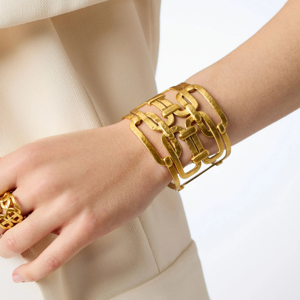A close-up of a person's wrist and hand wearing a chunky, multi-strand gold chain-link cuff bracelet and a matching gold ring, styled with a cream-colored blazer.