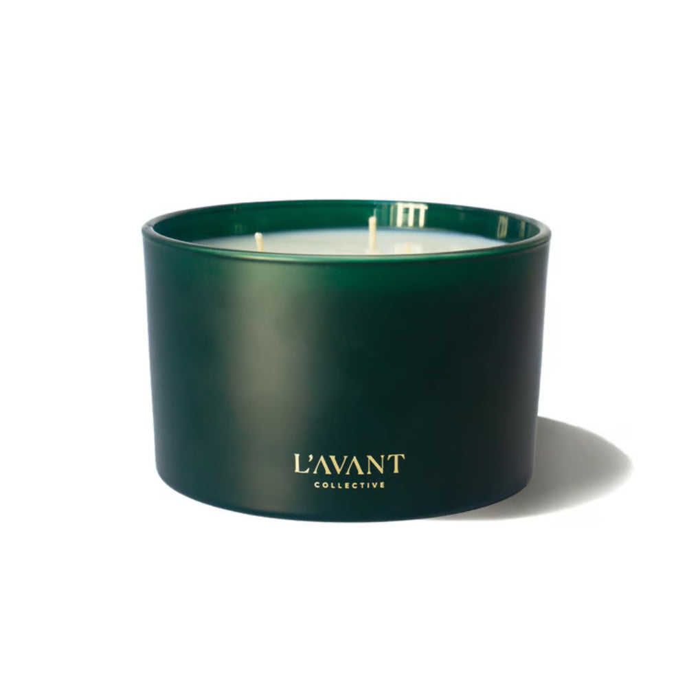A dark green glass jar candle with white wax and gold 'L'AVANT COLLECTIVE' branding, photographed on a clean white background with a soft shadow.