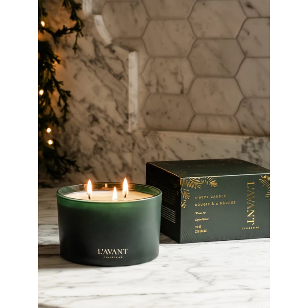 A dark green 3-wick candle in a matte jar with three lit flames, placed on a marble surface beside its black and gold L'AVANT COLLECTIONS box, with a softly blurred holiday scene and hexagonal tile wall in the background.