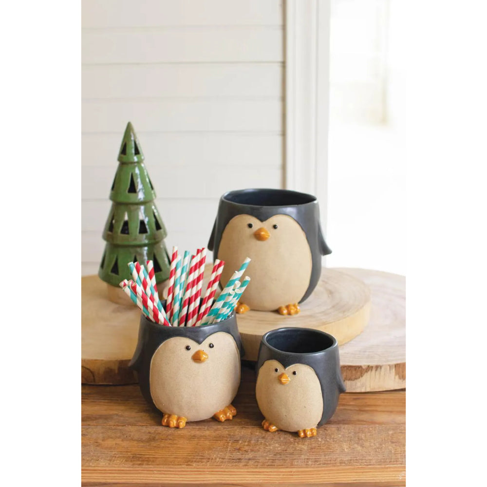 Three penguin-shaped ceramic mugs in dark gray and cream, arranged on a wooden surface with a small green tree decor piece and striped paper straws.