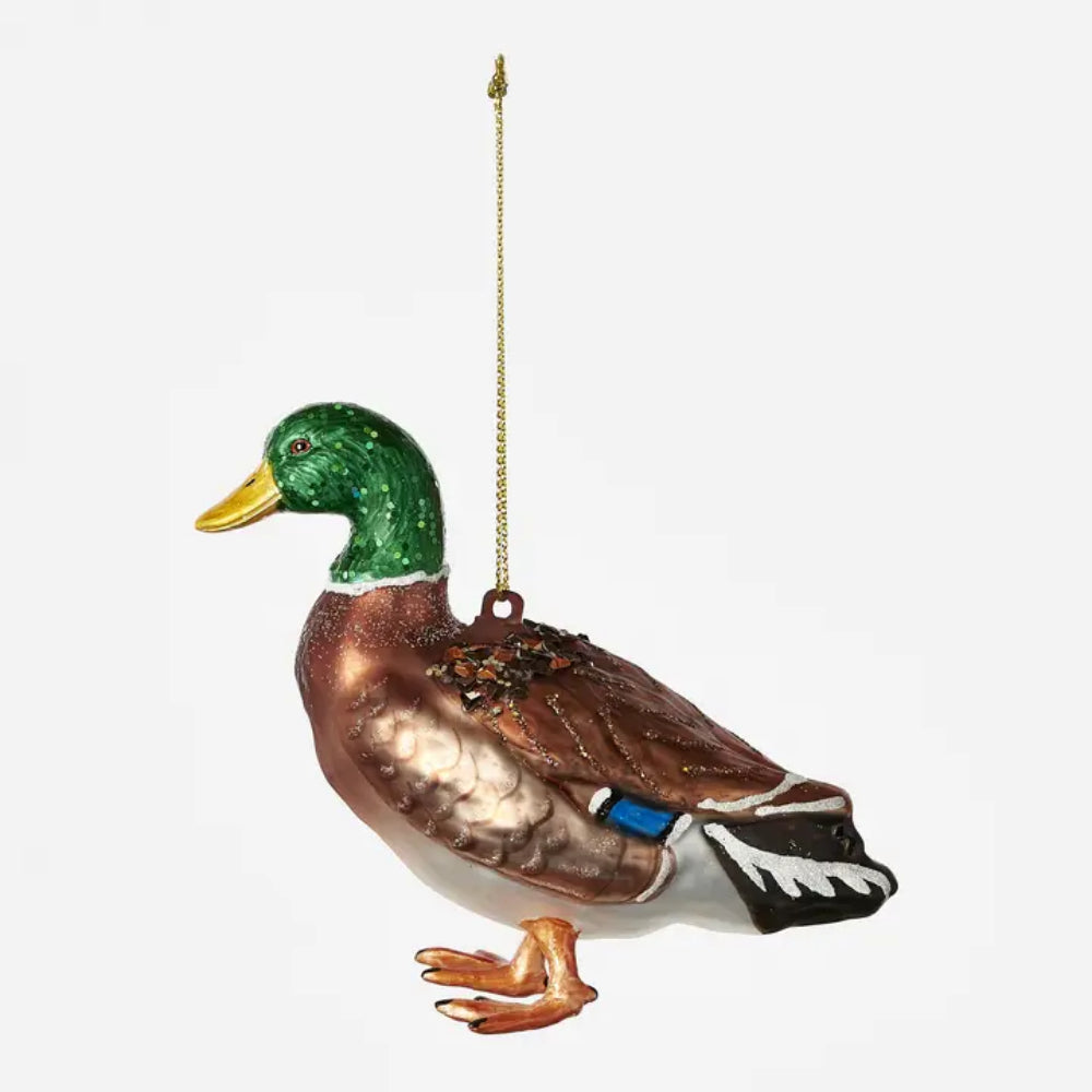 A decorative hanging ornament shaped like a mallard duck, shown against a plain light gray background. The ornament has a glossy green head, yellow beak, brown and copper-toned body, white underparts, black tail feathers with white tips, and a small blue accent near the tail.