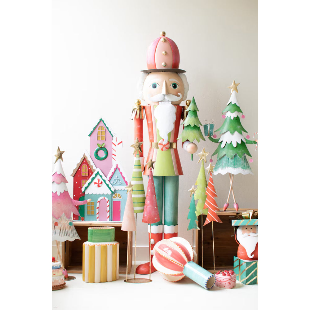 A festive holiday display featuring a large wooden nutcracker in the center, surrounded by colorful Christmas decorations including small paper trees, a house, ornaments, and gift boxes on a white background.