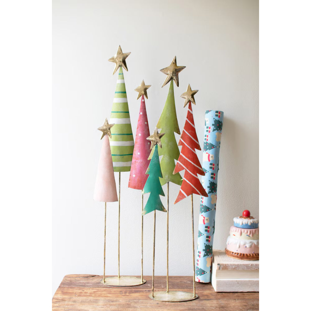 A set of decorative Christmas tree-shaped table centerpieces with gold star toppers, displayed on a wooden surface against a plain light wall. The trees come in pastel pink, green, teal, red, and blue patterned designs, with some standing upright and others rolled up.