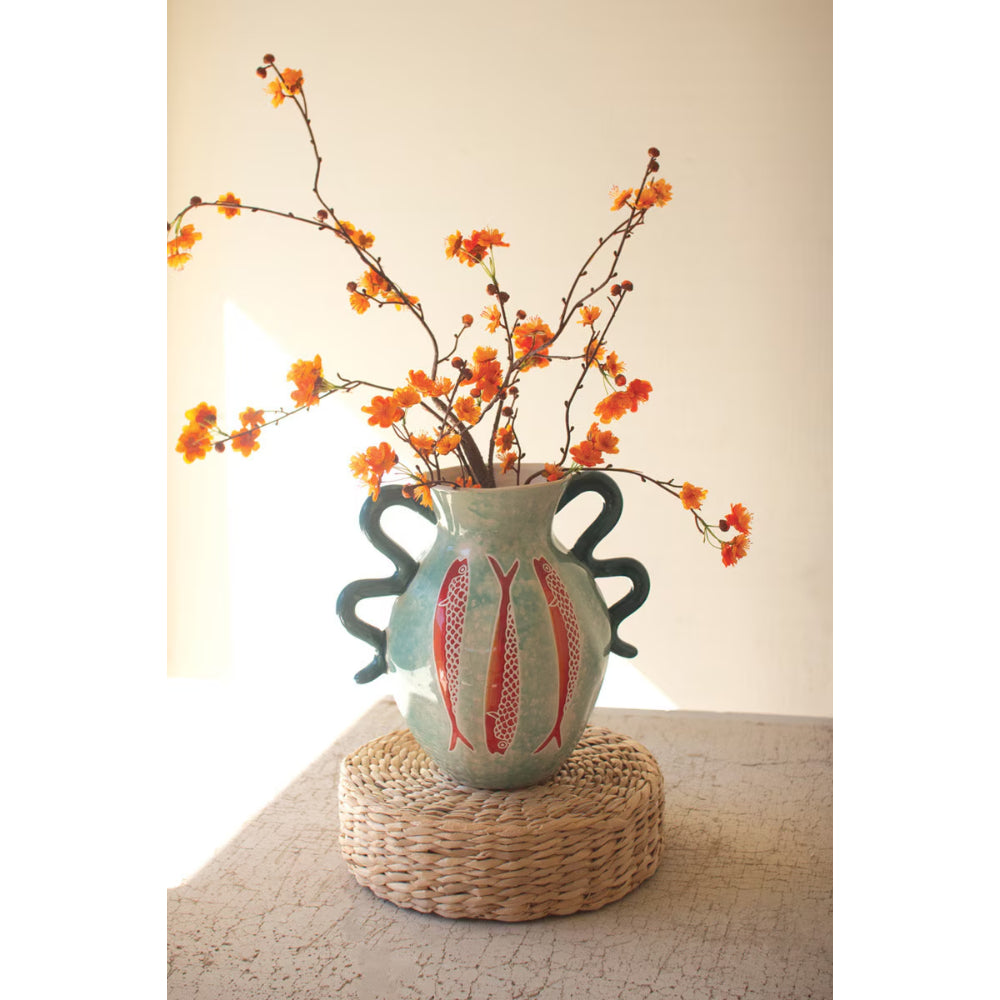 A handmade ceramic vase with three dark green handles and red fish motifs on a light green body, filled with orange-blossomed branches, displayed on a woven round mat.