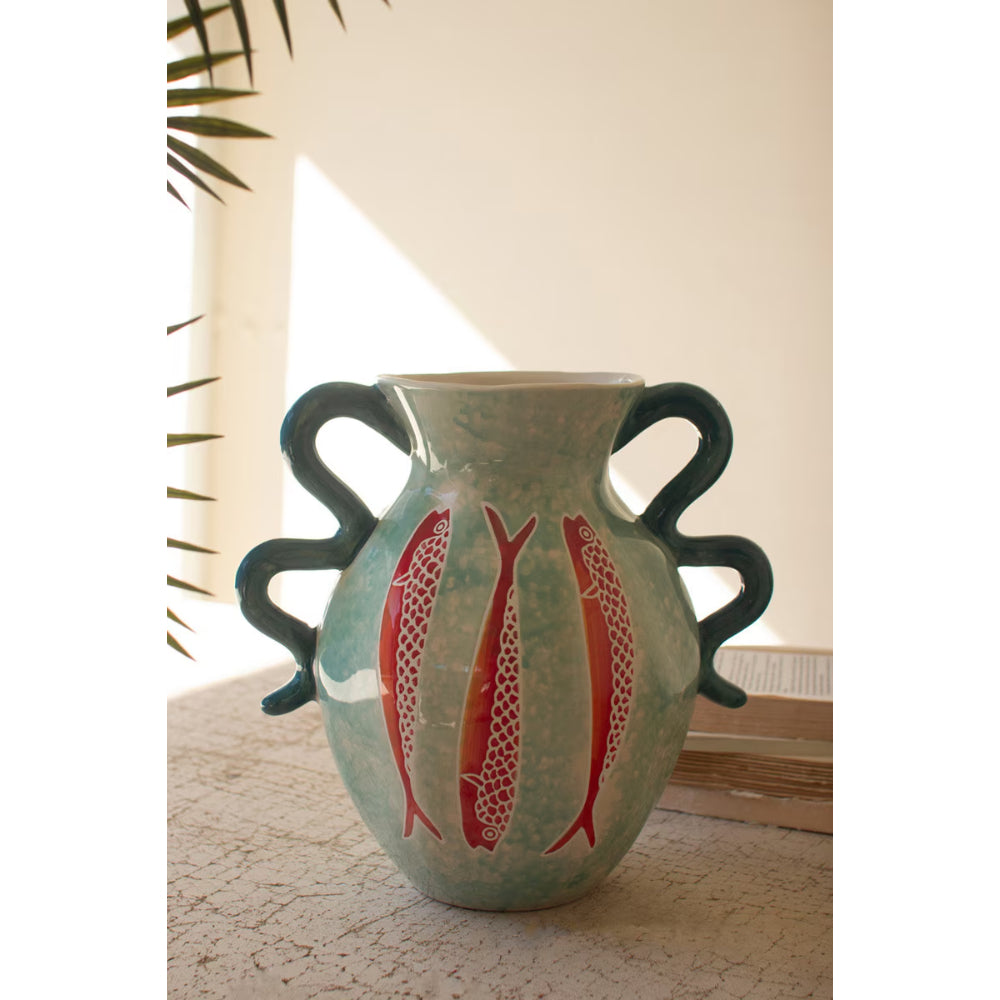 A handmade ceramic vase with a light green body, dark green handles, and red fish patterns, displayed on a textured surface with a warm neutral background.