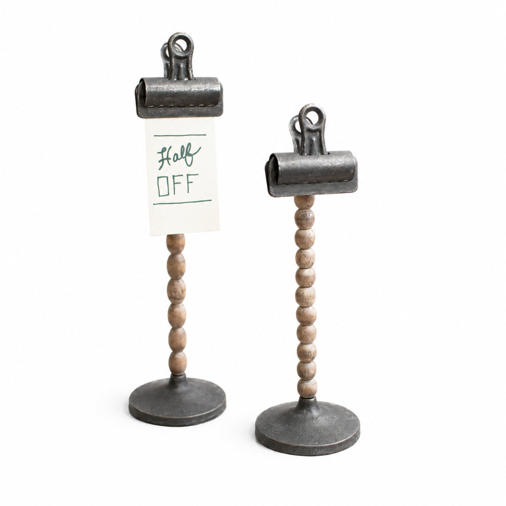 Two tabletop sign holders with dark metal clip tops, beaded wood stems, and round bases on a white background. One holder has a small white card attached that reads 'Half OFF'.
