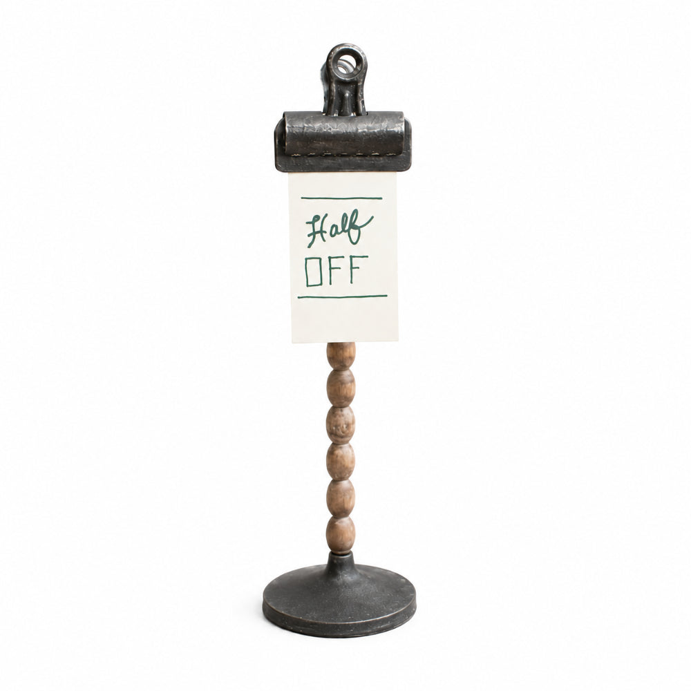 A tabletop sign holder with a round black metal base, a vertical beaded wood stem, and a black metal clip holding an off-white sign that reads 'Half OFF' in green handwriting.
