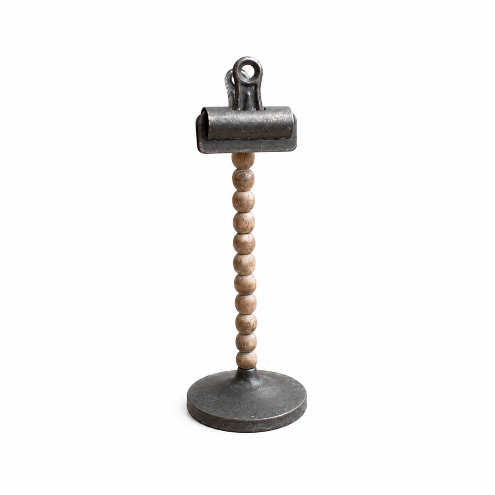 A decorative tabletop stand featuring a round dark metal base, a vertical stem made of small light wood beads, and a matching dark metal clip at the top, shown on a plain white background.