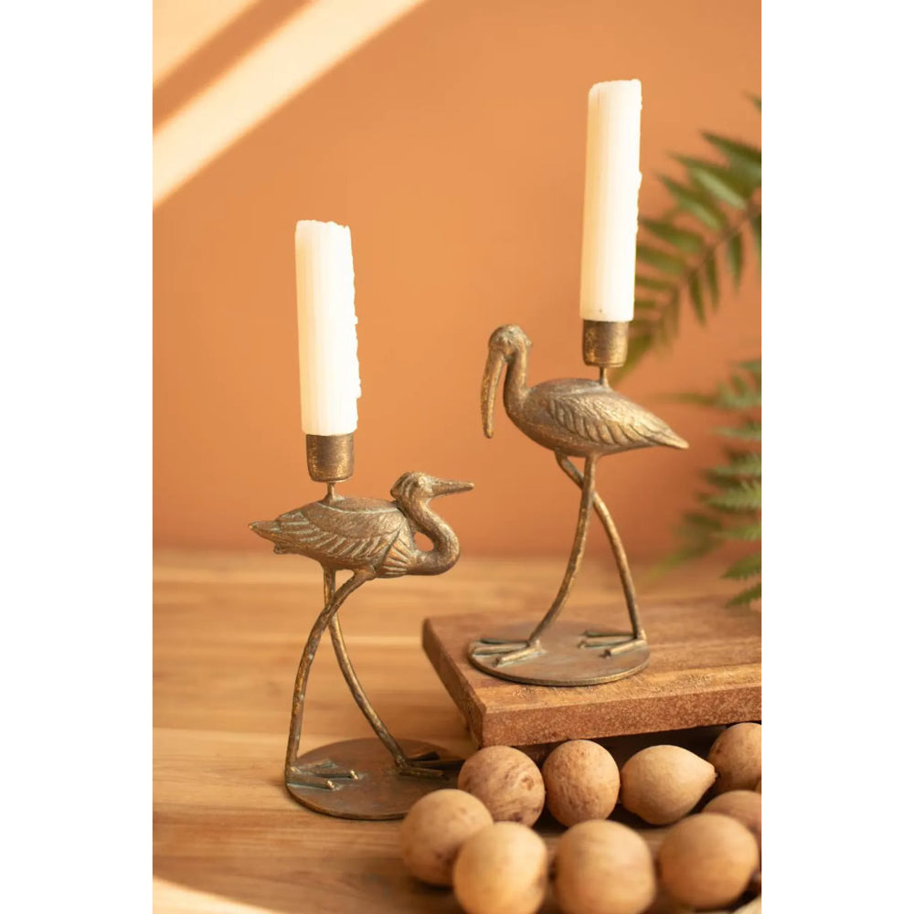 Two bronze-toned metal stork candle holders on a wooden surface, each holding a white taper candle, with a warm orange background and decorative props including nuts and a plant.