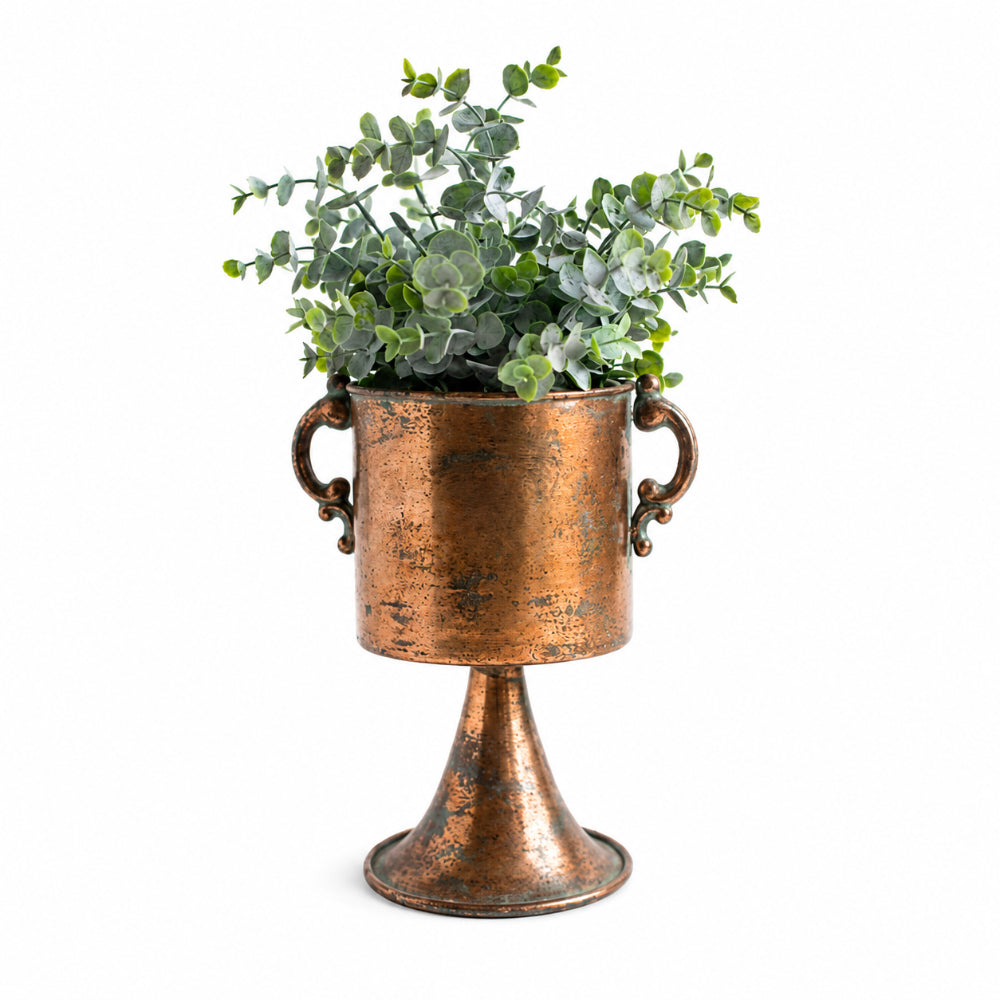 A distressed copper pedestal planter with side handles, filled with green eucalyptus foliage, set against a plain white background.