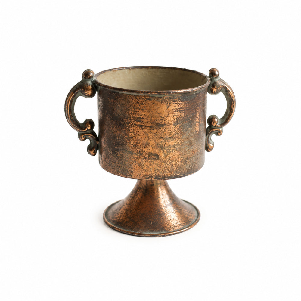 A small decorative cup or goblet with a pedestal base and two ornate side handles, shown on a plain white background. The finish is distressed metallic copper/bronze with darker patina details.