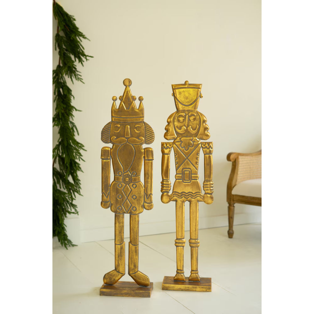Two gold nutcrackers standing on a white floor with a decorative plant and chair in the background.