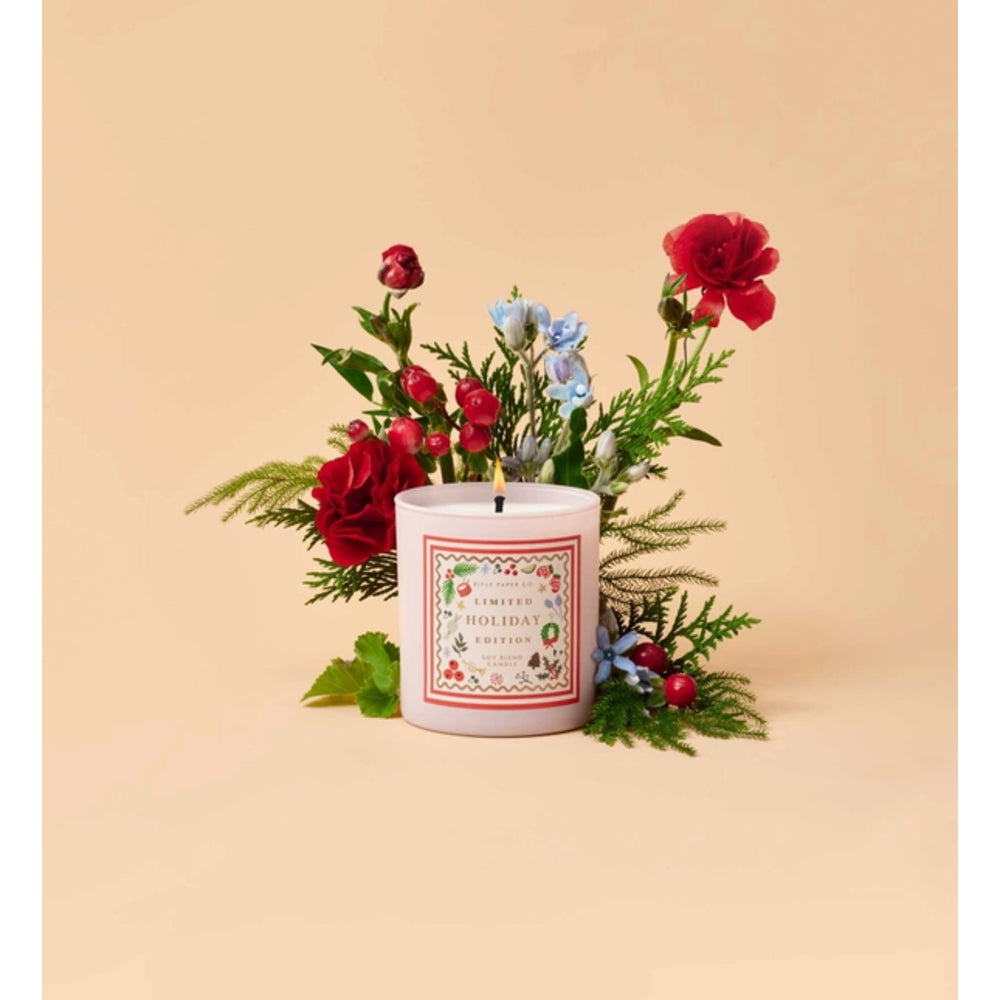 A white candle with a decorative holiday-themed label sits in front of a floral arrangement of red roses, blue flowers, and green foliage on a warm beige background.