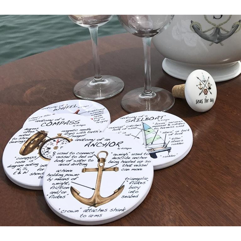 Four round white coasters with nautical-themed printed designs and one matching white wine stopper, arranged on a wooden table with wine glasses and a ceramic container in the background.
