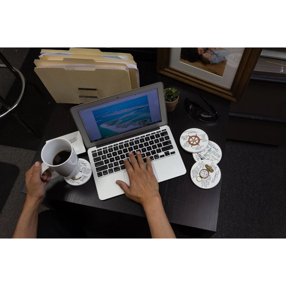Overhead view of a person using a silver laptop at a dark desk, holding a white mug on a saucer. The desk also holds a pair of sunglasses, a small plant, framed photos, and a stack of files.