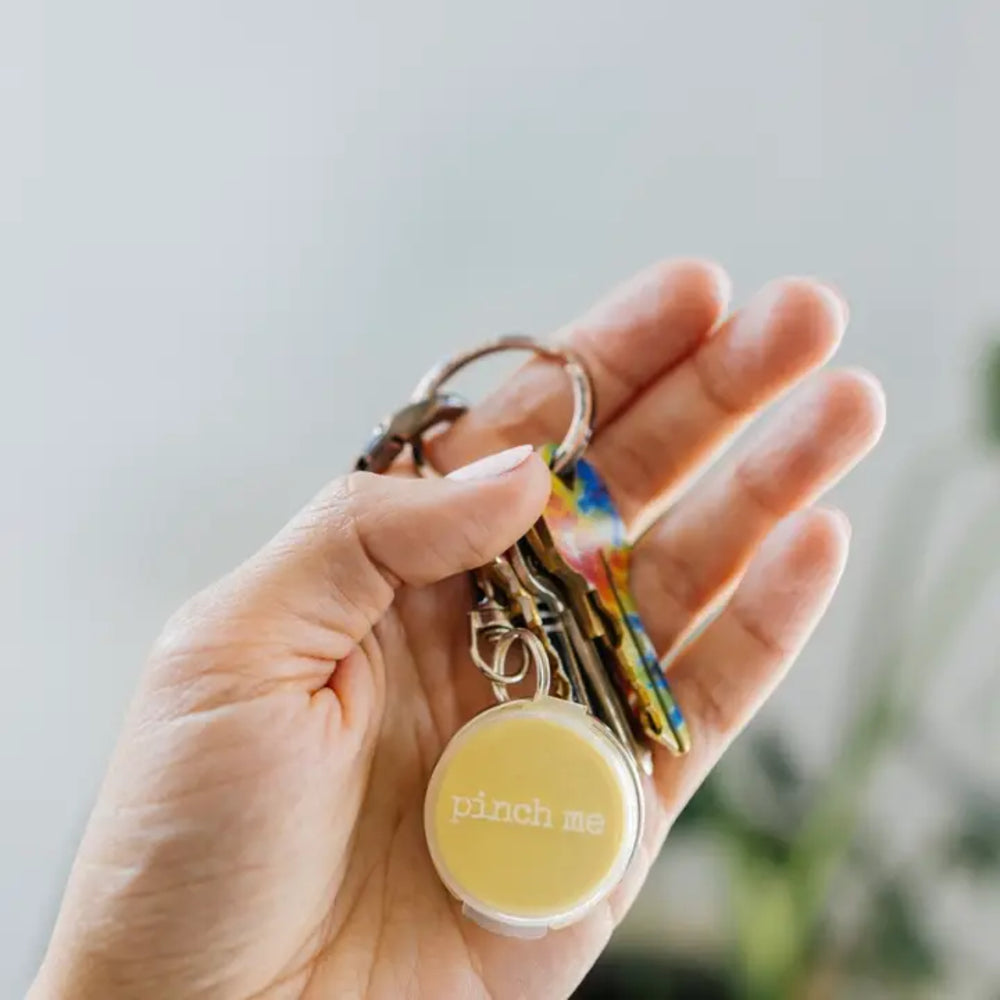 A hand holding a keyring with a round pale yellow container that reads 'pinch me' in white text, alongside several keys.