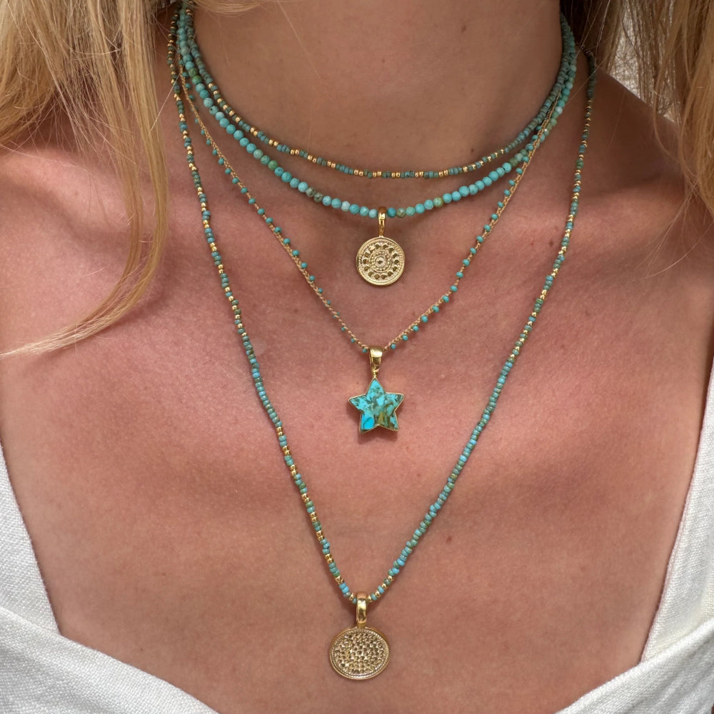 Close up image of a woman with blonde hair wearing a stack of turquoise and gold beaded necklaces with different pendants.