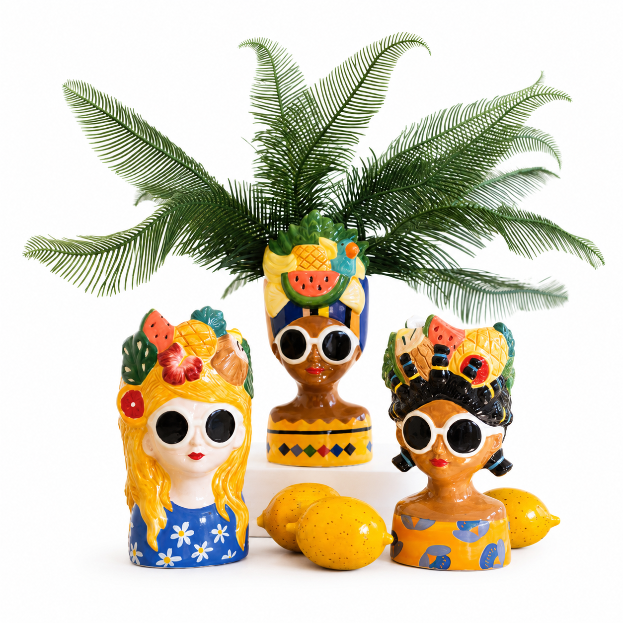 Three colorful decorative figurines shaped like stylized women with large black sunglasses, floral and fruit headdresses, and yellow dresses, arranged with two lemons and a green palm frond on a white background.