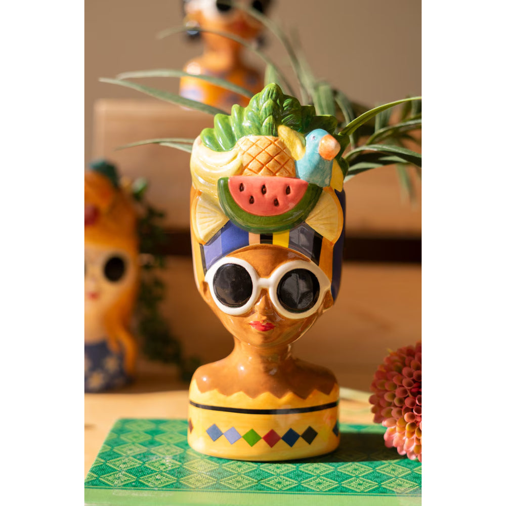 Colorful ceramic vase shaped like a stylized face with a pineapple on its head, placed on a green surface.