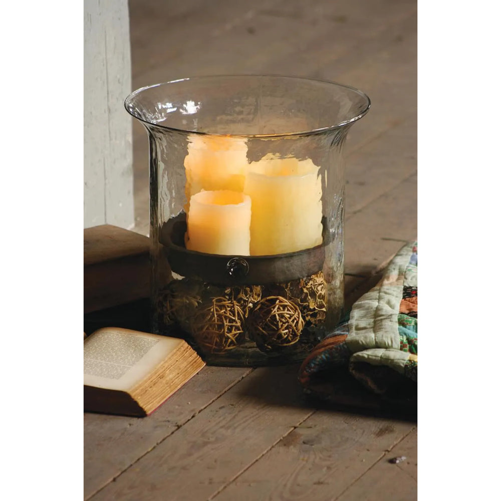 A clear glass candle holder with a flared rim, containing two lit cream pillar candles and decorative woven balls, styled on a rustic wooden floor with an open book and quilt nearby.