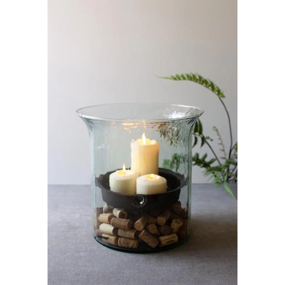 A clear glass vase or candle holder filled with brown wine corks at the bottom, holding a black candle holder with three lit white pillar candles, set on a gray surface against a light gray background with a blurred green plant.