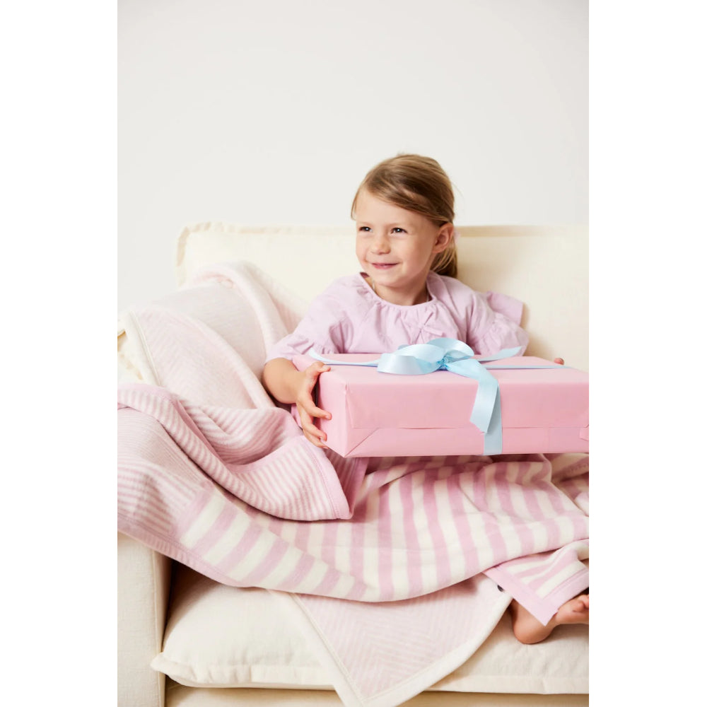 Child sitting on a couch holding a pink gift box with a blue ribbon with pink and white striped mini blanket. 