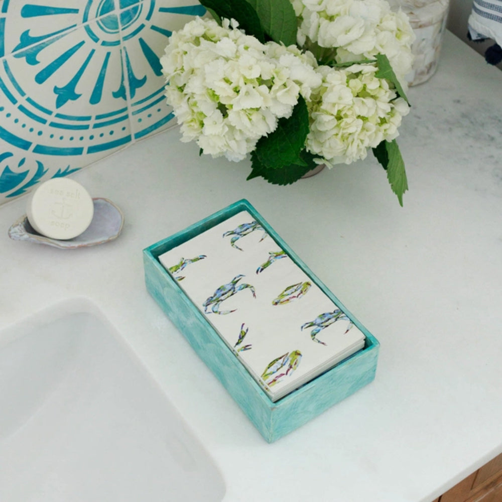 Teal box with patterned towels on a white countertop with flowers and soap.