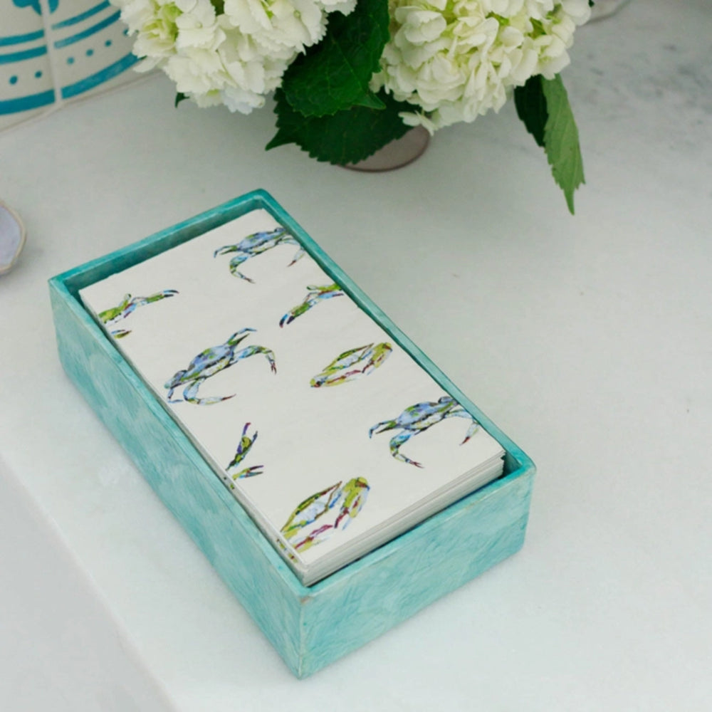 Stack of napkins with crab pattern in a turquoise holder on a white surface with flowers in the background