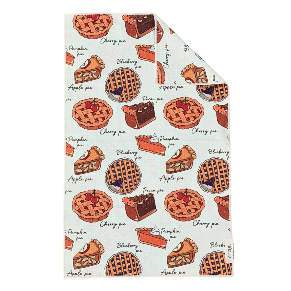 Hand towel featuring a repeating pattern of illustrated pies and cake slices with labels such as cherry pie, pumpkin pie, apple pie, and chocolate pie. The top-right corner is folded over to show the fabric texture and reverse side, set against a plain white background.