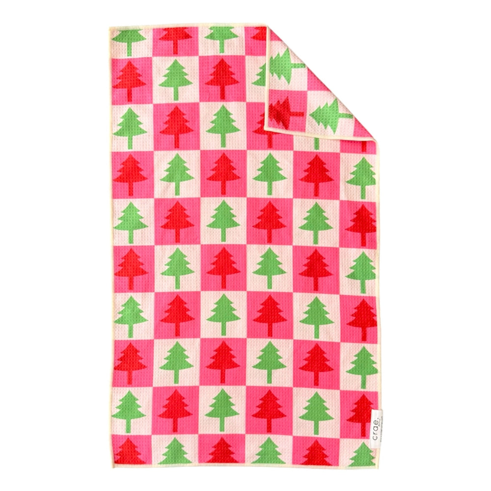 A square kitchen towel with a festive Christmas tree checkerboard pattern in red and green on a pink and cream background, shown with one corner folded over on a white background.