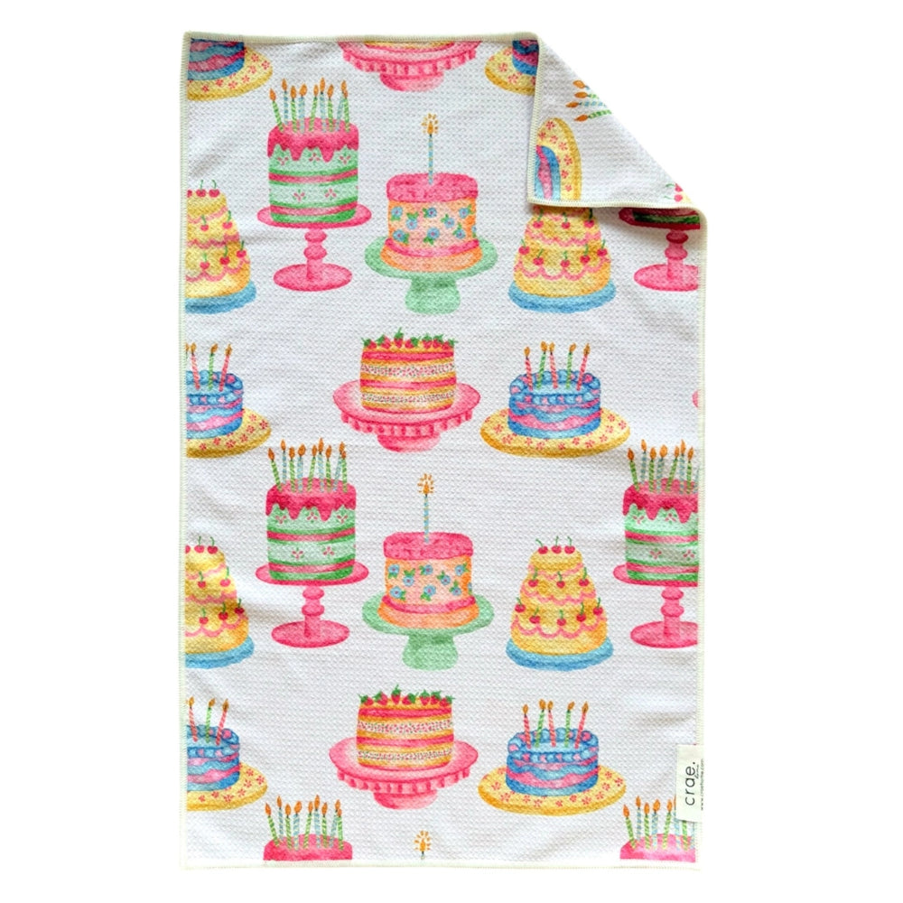 A grey hand towel with birthday cake designs on a white backround. 