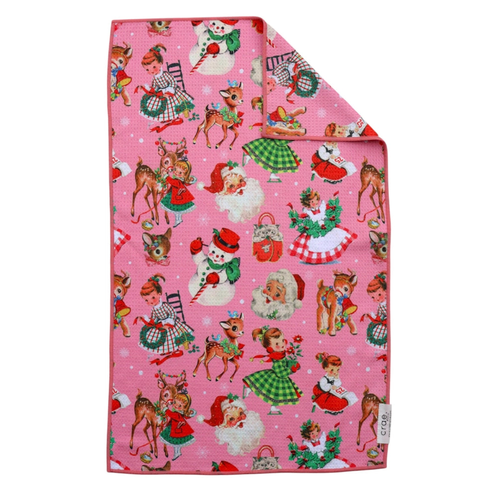A pink fabric item, likely a towel or blanket, featuring a vintage Christmas print with Santa Claus, snowman, elves, and deer. The top-right corner is folded over to show the reverse side, which is solid pink.