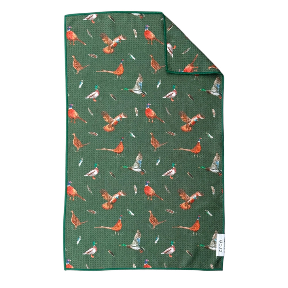 A green woven towel with a repeating pattern of pheasant birds and feathers, featuring a solid green border and a small white brand tag on the bottom right corner.
