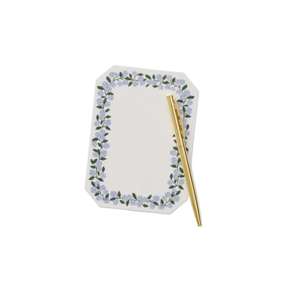 A photo of a note pad and gold pen of a  note writing set by Rifle Paper Co. featuring hydrangea illustrations, including 130 flat notes and a gold pen on a white backround. 