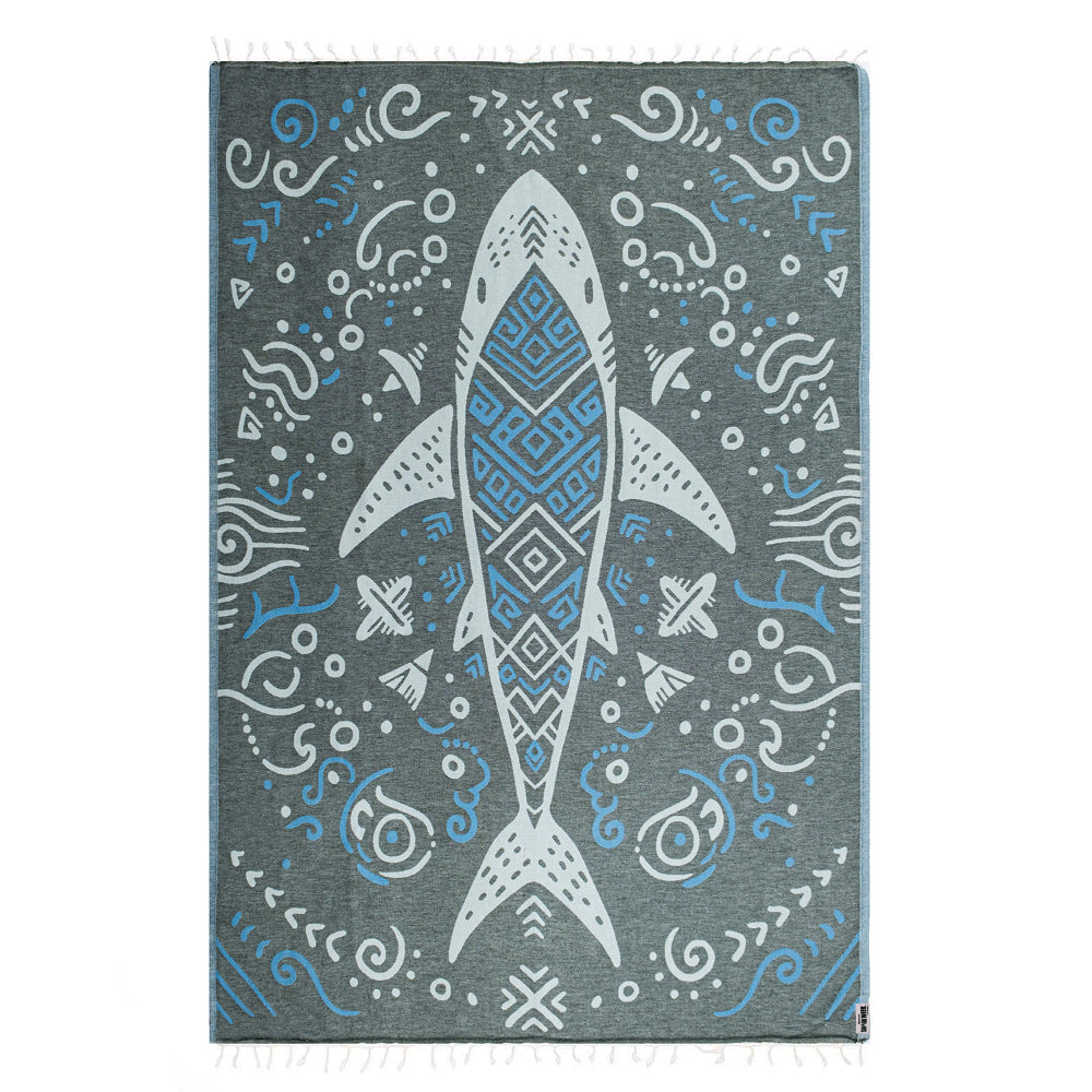 Side one of a blue & gray beach towel with a shark design with blue abstract patterns and a white tassel edge on a white background.