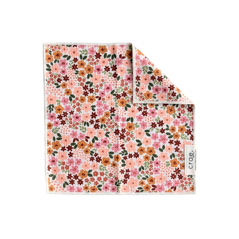 A square cotton wash cloth featuring a dense floral print in pink, orange, brown, and green on a light pink background. The pocket square is shown folded with one corner turned over to reveal the reverse side, which has a white tag with the brand name 'crae'.