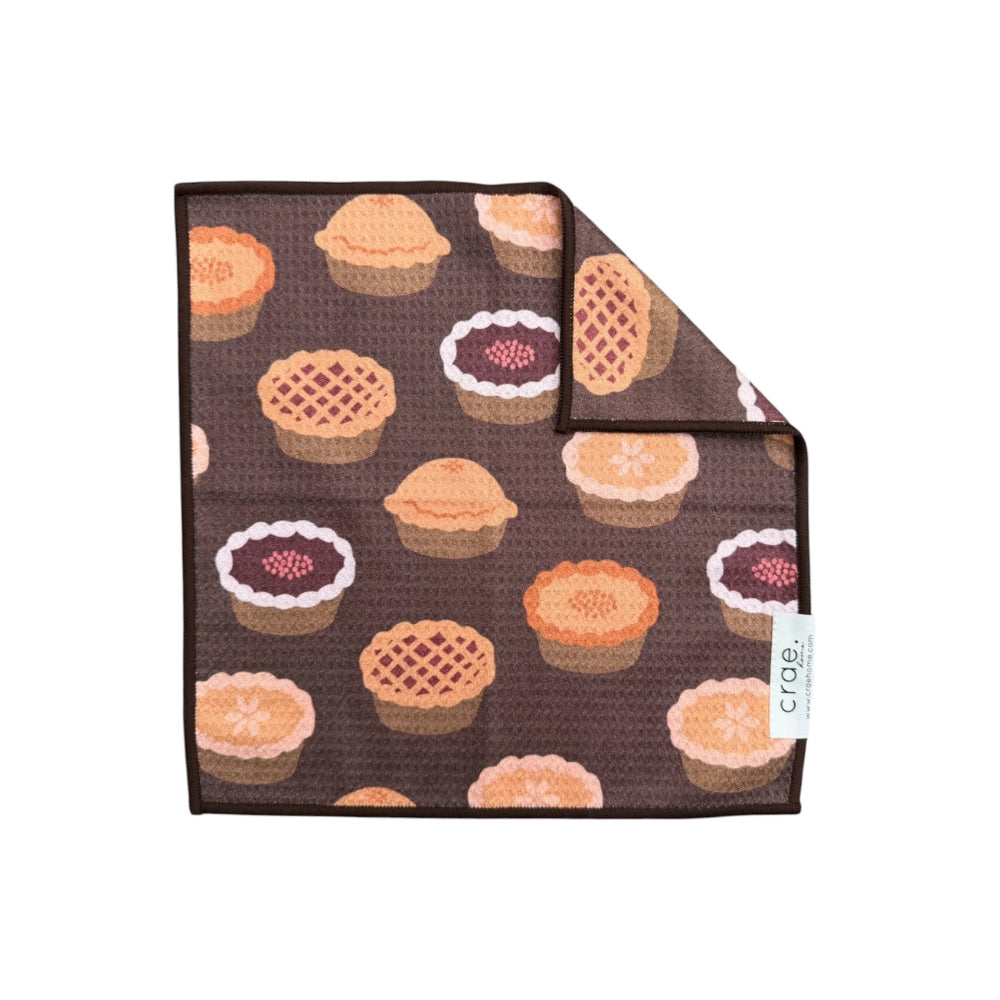 A square wash cloth with brown background and a repeating pattern of assorted pies and tarts. One corner is folded over to show the reverse side, which is plain brown.