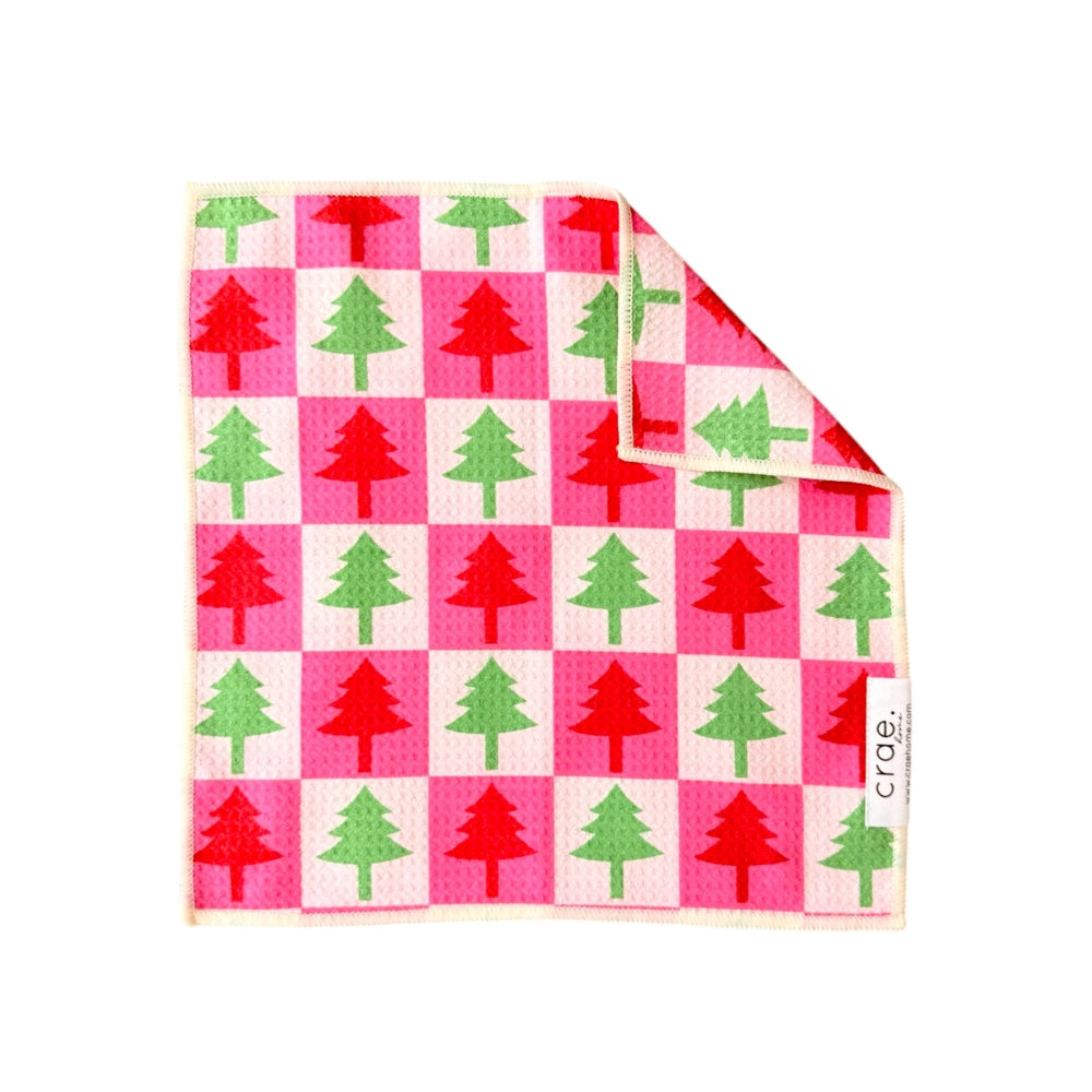 A square washcloth featuring a pink and white checkered pattern with green and red Christmas tree motifs. The top-right corner is folded over to show the reverse side, which has a white brand label reading 'Crae.