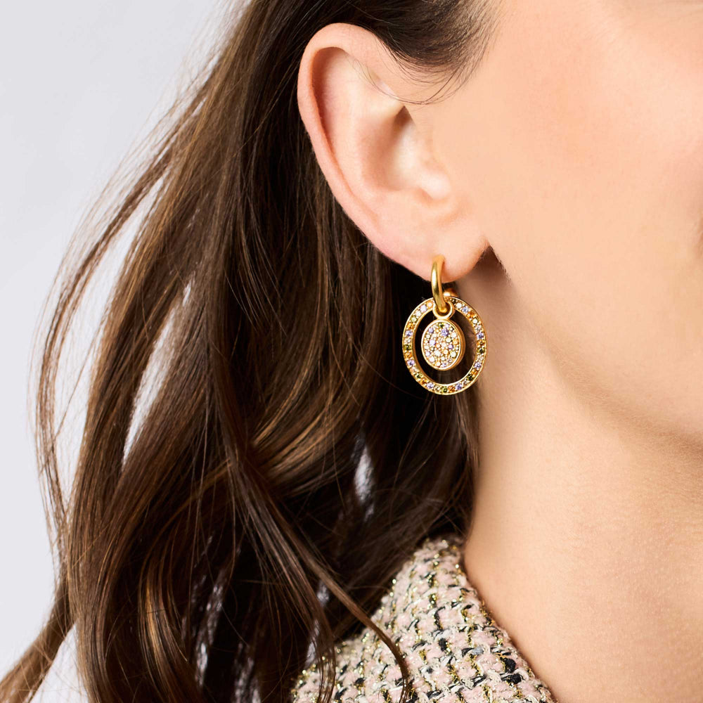Close-up of a person wearing a gold earring with a clear gemstone on a neutral background