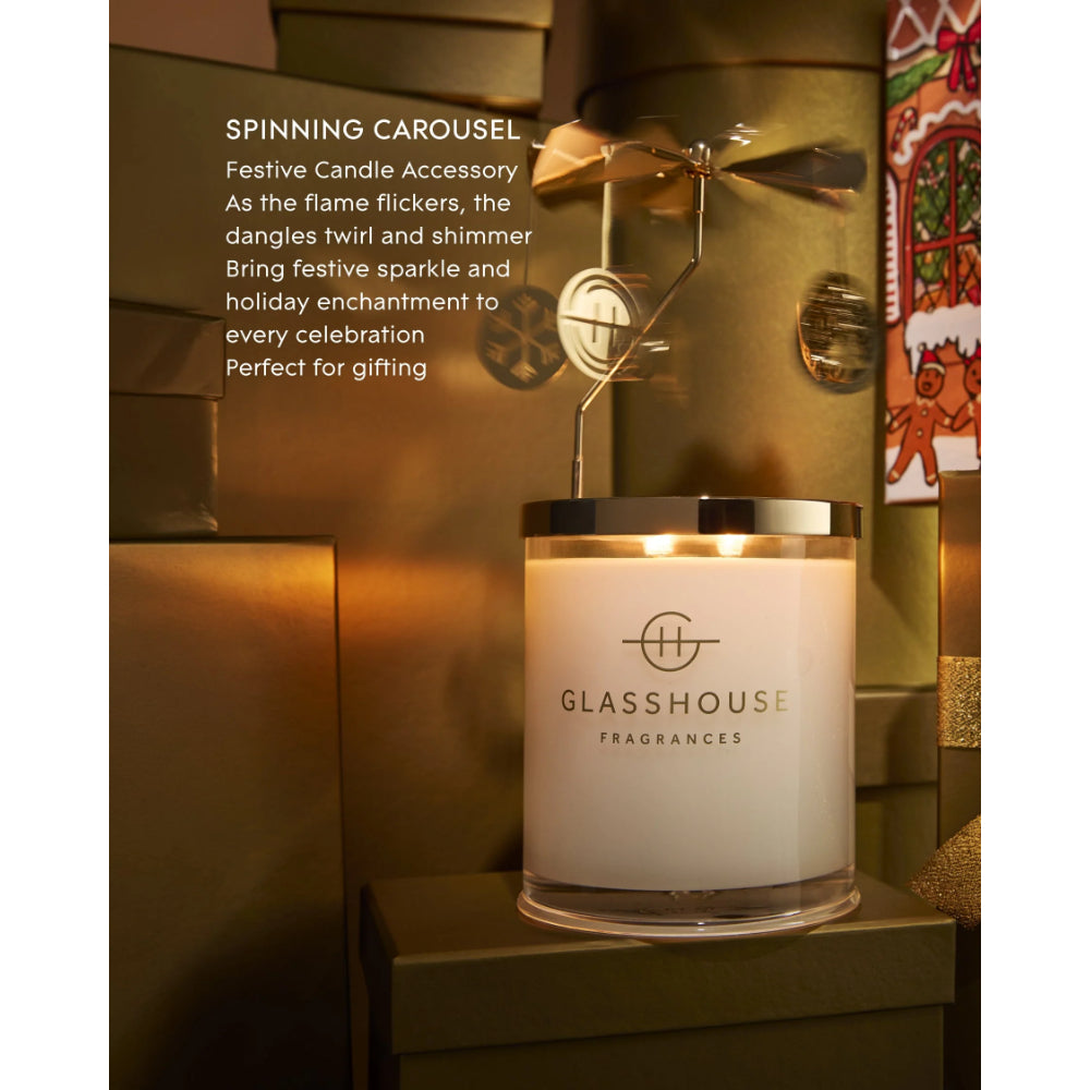 A lit Glasshouse Fragrances candle in a clear glass jar with a silver lid, placed on a dark pedestal in a warm holiday-themed setting. White text on the left reads 'SPINNING CAROUSEL' with descriptive copy about the candle accessory.