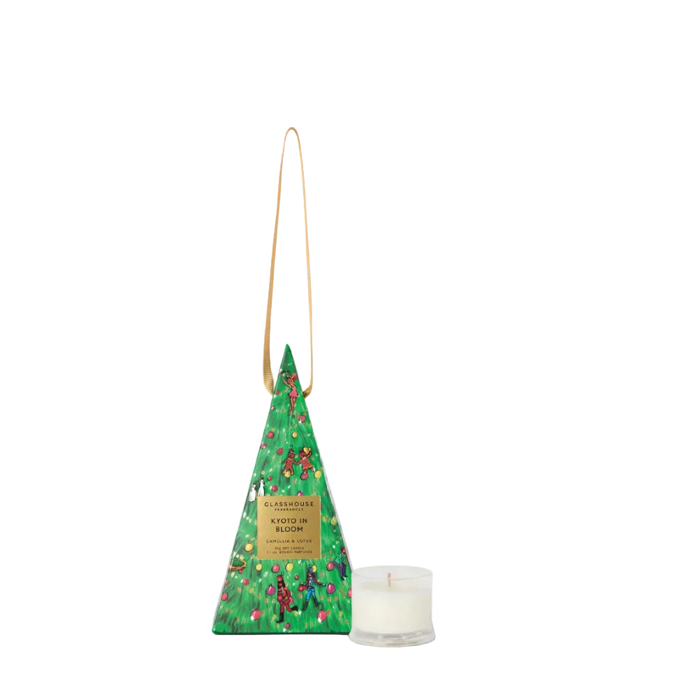 A Christmas-themed candle in a triangular green gift box with a gold hanging loop, shown next to a small white candle in a clear glass holder on a light gray background.